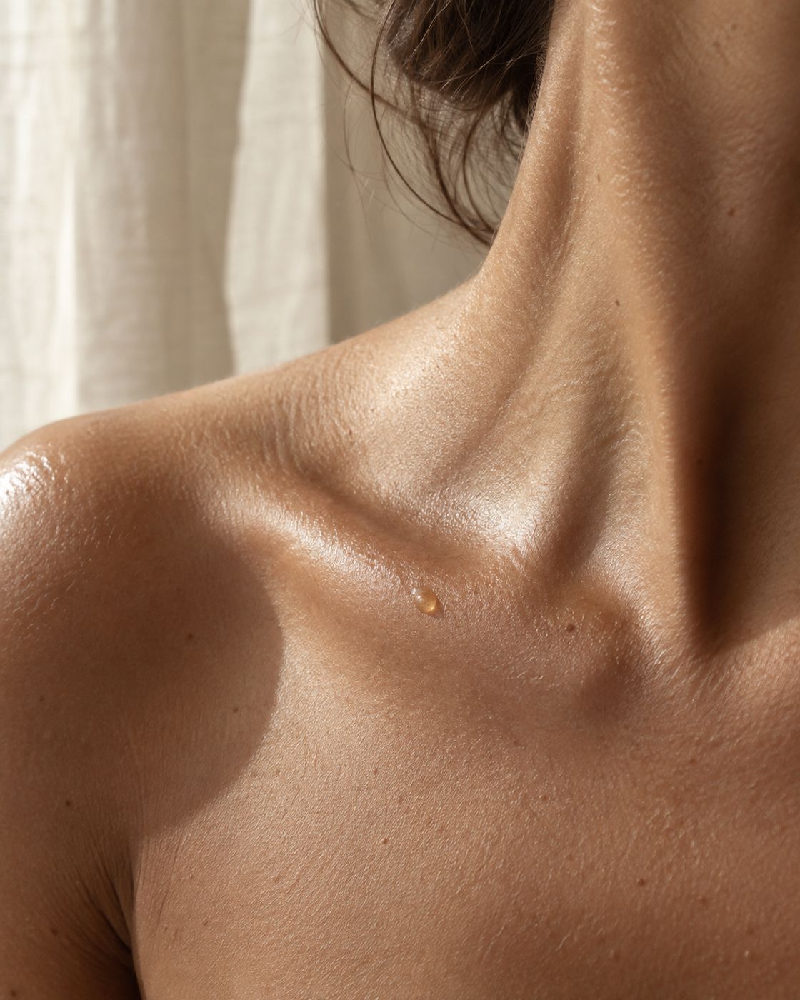 Close crop of a bare shoulder and collarbone in soft morning light, with a drop of golden oil on the skin.